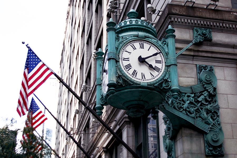 About Our Agency - Old Green Ornate Metal Clock Mounted on the Corner of a Stone Building in Chicago, With Flagpoles Attached to the Building Waving American Flags